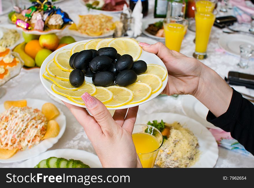 Black olives and lemons on a plate in female hands over a celebratory table. Black olives and lemons on a plate in female hands over a celebratory table.