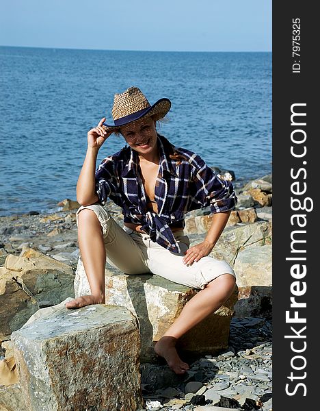 The beautiful woman in a hat sits on a stone beach. The beautiful woman in a hat sits on a stone beach