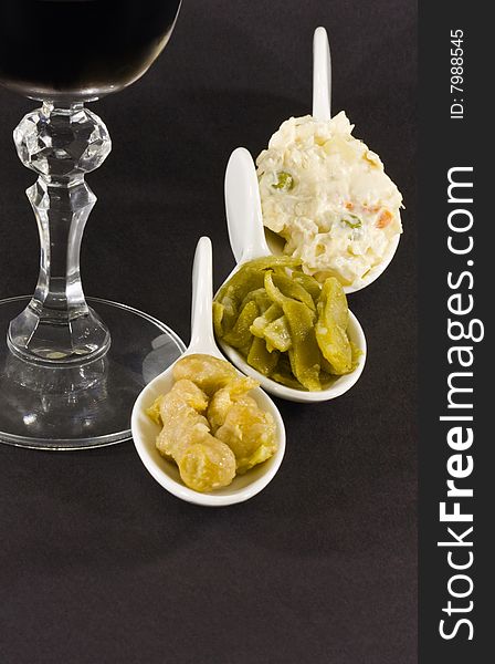 Russian salad, chick-peas and cases in white spoons and wine glass on black background