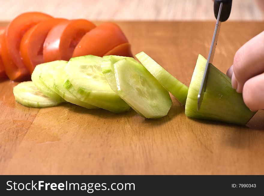 Hands slicing cucumber  on wooden board. Tomatoe in background.