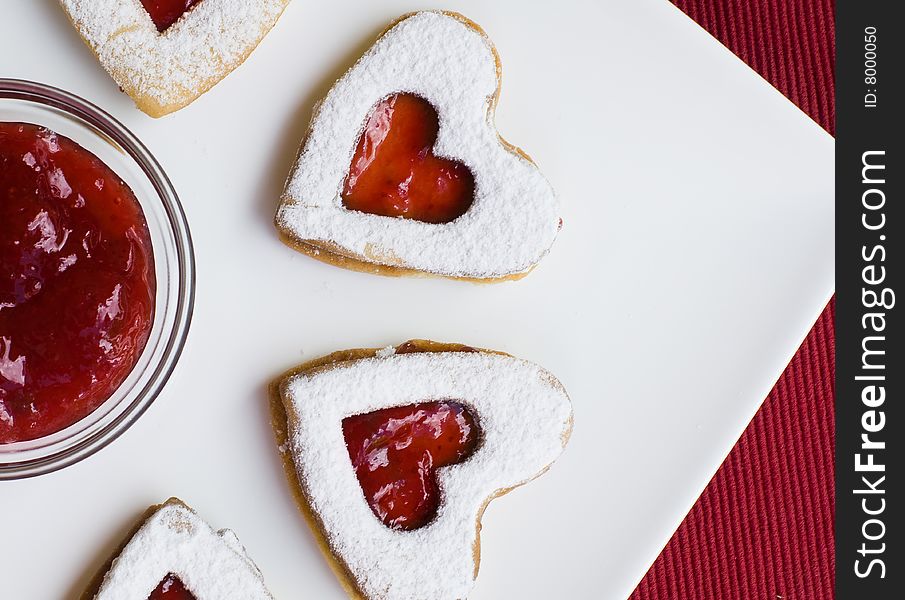 Heart shaped homemade jam cookies placed on a white plate with jam and a red background. Heart shaped homemade jam cookies placed on a white plate with jam and a red background
