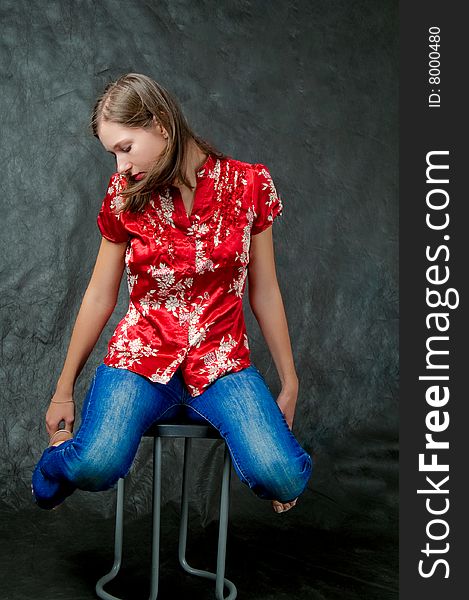 Young dark haired girl sitting on chair with legs lifted up and hands touching hips looking down thinking isolated on black background