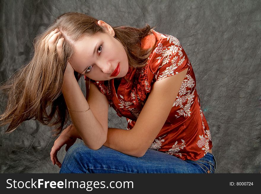 Pretty young girl sitting turned with hand lifted and touching hair legs crossed appear looking forward thinking isolated on gray background. Pretty young girl sitting turned with hand lifted and touching hair legs crossed appear looking forward thinking isolated on gray background