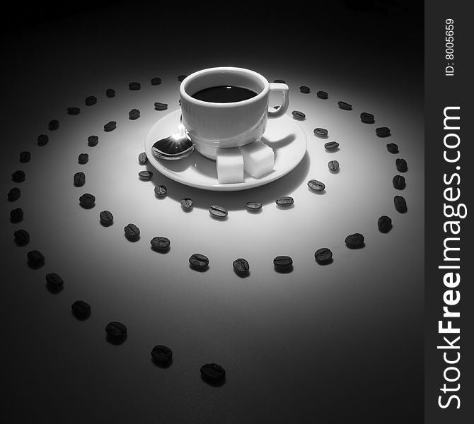 In the spotlight the cup of coffee on the saucer with two slices of sugar and spoon, at the center of spiral made of coffee grains. In the spotlight the cup of coffee on the saucer with two slices of sugar and spoon, at the center of spiral made of coffee grains.