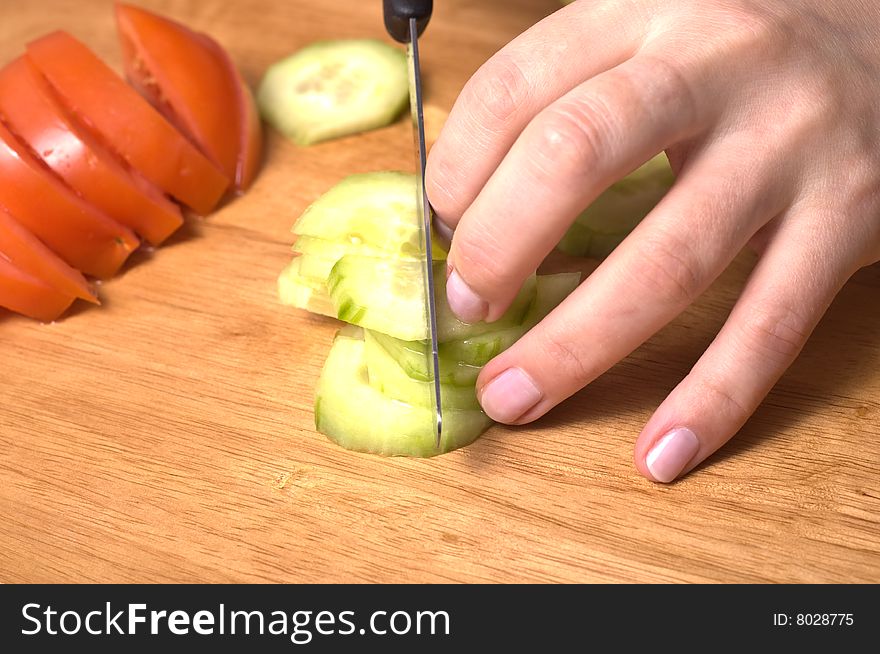 Hands slicing cucumber  on wooden board. Tomatoe in background.