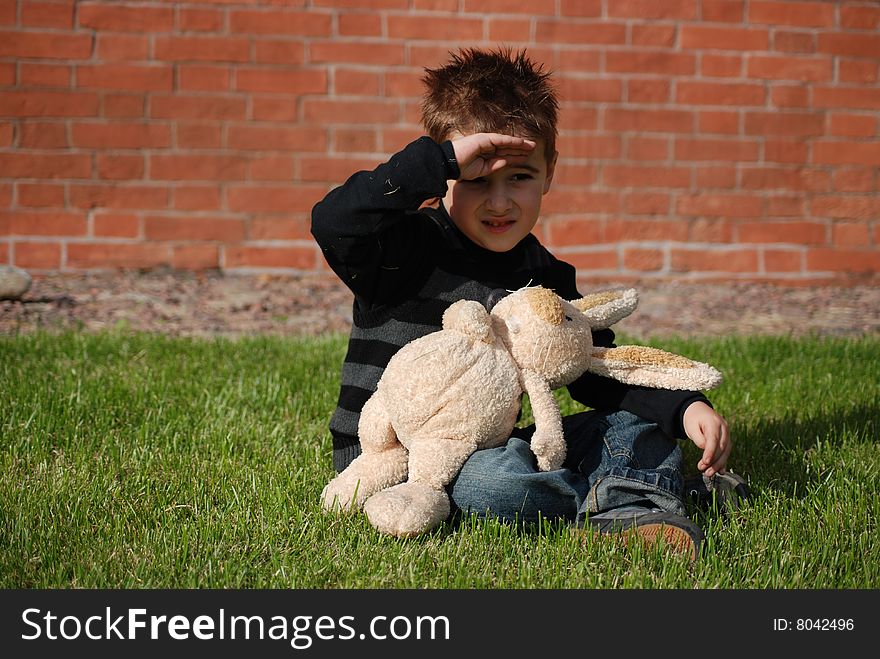 Little boy with a toy hare in hands, sitting on a green grass on a lawn on a background a brick wall. Little boy with a toy hare in hands, sitting on a green grass on a lawn on a background a brick wall