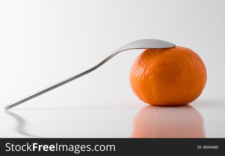 Metal teaspoon leaning on a tangerine, grey background, reflective surface