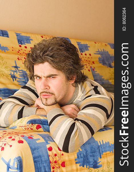 Young smiley dark haired caucasian man in striped sweater lying on country style sofa and frowning. Young smiley dark haired caucasian man in striped sweater lying on country style sofa and frowning