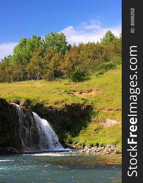 A small waterfall during summer, set against gentle hills with small trees on top of it. A small waterfall during summer, set against gentle hills with small trees on top of it