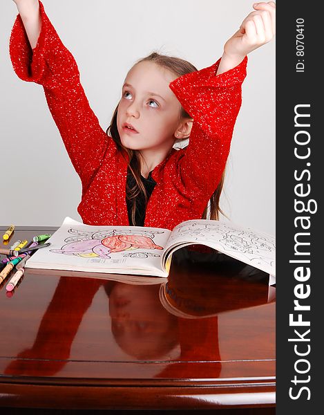 Little girl in a red sweater sitting on the table and coloring an book with crayons. Her face mirroring in the table and the arms are raised. Little girl in a red sweater sitting on the table and coloring an book with crayons. Her face mirroring in the table and the arms are raised.