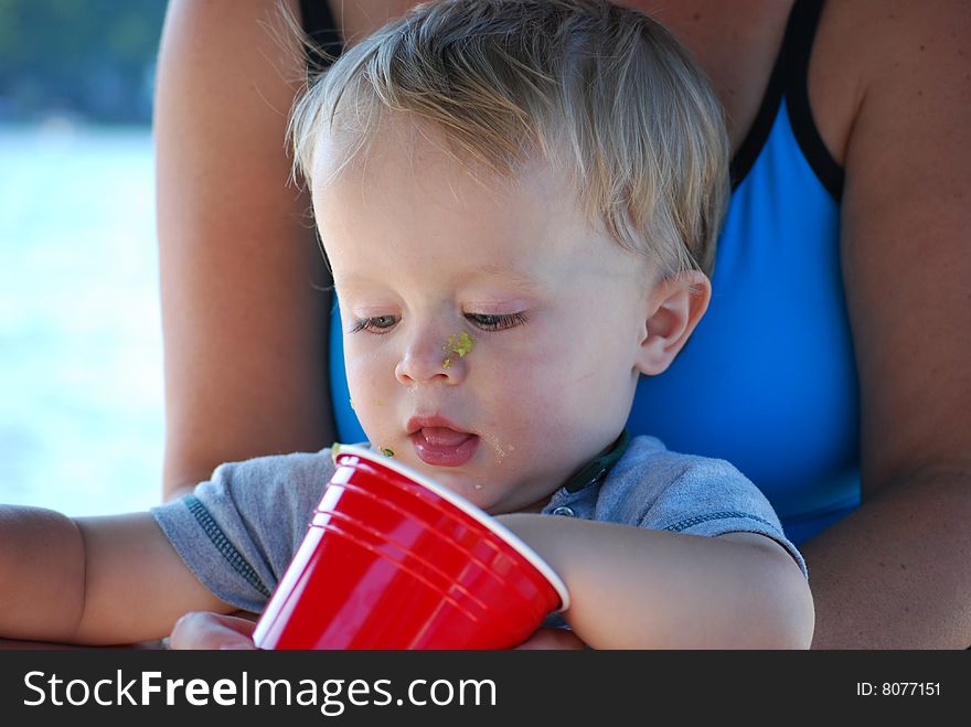 A messy baby eat guacamole from a cup while on his mother's lap at the beach. A messy baby eat guacamole from a cup while on his mother's lap at the beach