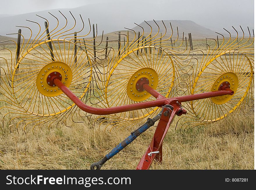 The tines of a large red, yellow, and blue farm implement dominate the foreground of a misty Montana landscape in autumn. Yellow grasses and barbed wire fence stretch out across a cattle ranch toward the mist-enshrouded foothills of the Front Range, near Choteau, Montana. The tines of a large red, yellow, and blue farm implement dominate the foreground of a misty Montana landscape in autumn. Yellow grasses and barbed wire fence stretch out across a cattle ranch toward the mist-enshrouded foothills of the Front Range, near Choteau, Montana.