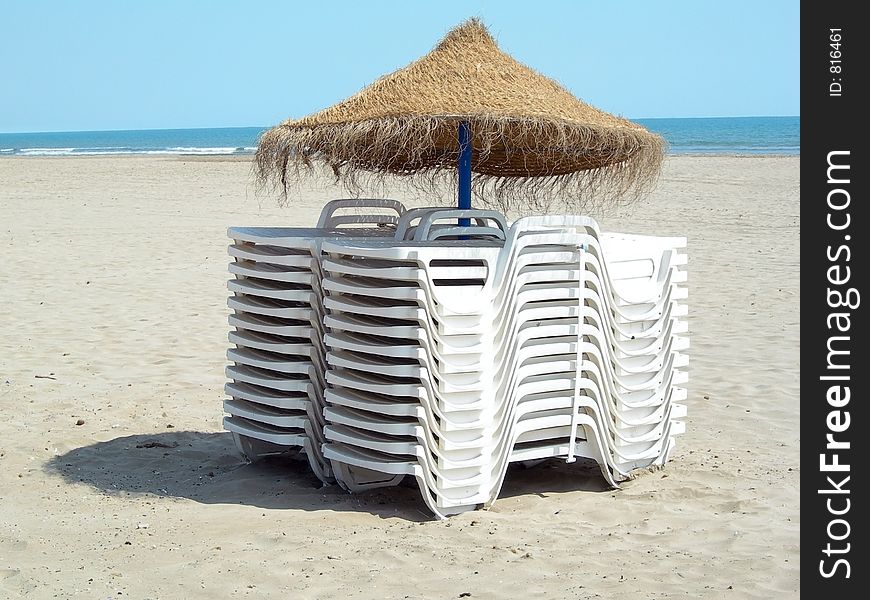 Two piles of white beach chairs under an umbrella in the sand. Two piles of white beach chairs under an umbrella in the sand