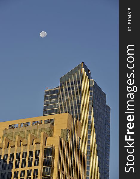 The moon in a blue sky at sunrise, over buildings, one skyscraper with glass. The moon in a blue sky at sunrise, over buildings, one skyscraper with glass.