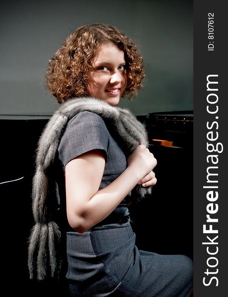 Pretty young dark haired girl with curly hair sitting turned near piano in grey dress with fur scarf smiling isolated