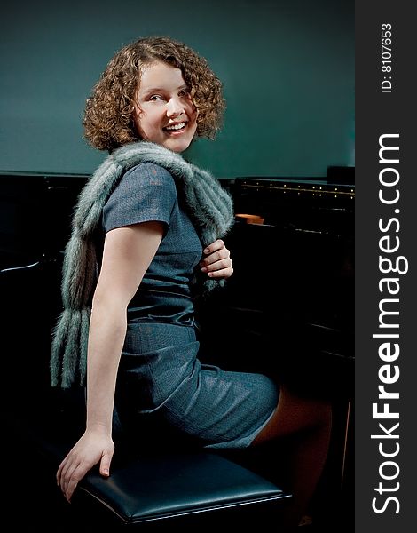 Pretty young dark haired girl with curly hair sitting turned near piano smiling in grey dress with fur scarf smiling isolated