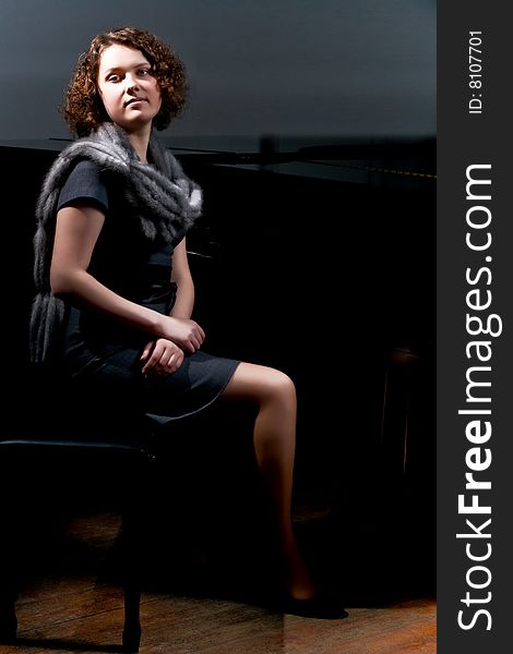 Pretty young lady sitting near black piano in grey dress with fur scarf around neck isolated