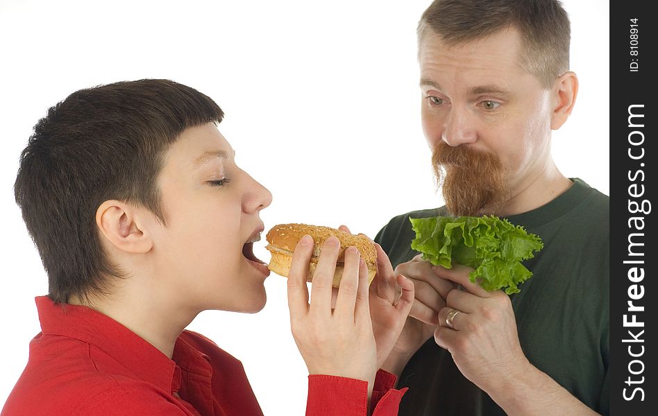 A breaded man with lettuce in hands watching enviously at a woman who is going to bite a hamburger. A breaded man with lettuce in hands watching enviously at a woman who is going to bite a hamburger
