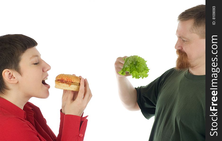 A woman is about to bite a hamburger while a breaded man standing beside her stares in surprise at lettuce leaves in his hand. A woman is about to bite a hamburger while a breaded man standing beside her stares in surprise at lettuce leaves in his hand