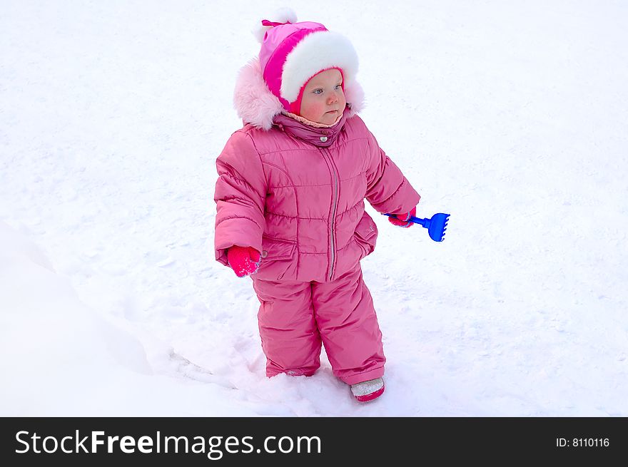 Pretty little girl with blue plastic toy rake in winter outerwear stay on snow-covered ground. Pretty little girl with blue plastic toy rake in winter outerwear stay on snow-covered ground.