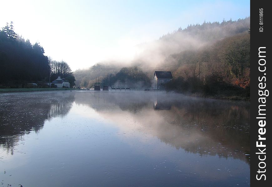 A white building stands beside a still lake whose waters reflect the tree covered hills and mist. A white building stands beside a still lake whose waters reflect the tree covered hills and mist