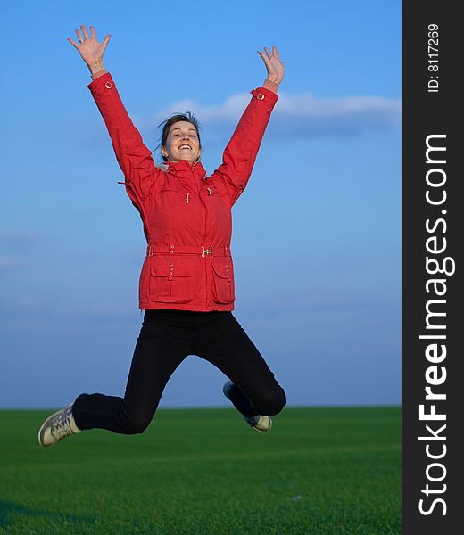 Jumping girl in red jacket on blue sky and green grass background