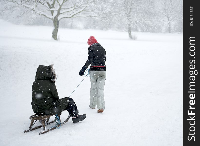 The bare branches of Winter trees are caked in heavy snow. In the foreground a girl is seen pulling her friend on a sledge. Both figures have their backs to the camera and their faces are hidden. The bare branches of Winter trees are caked in heavy snow. In the foreground a girl is seen pulling her friend on a sledge. Both figures have their backs to the camera and their faces are hidden