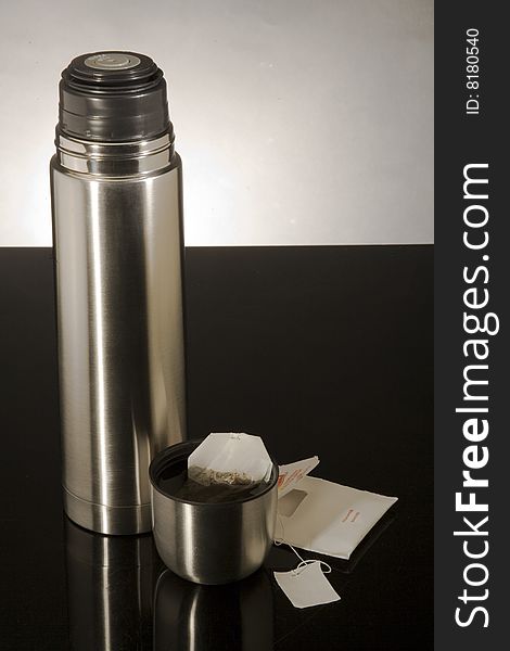 Metal flask with tea bag on a black reflective surface against a gray background