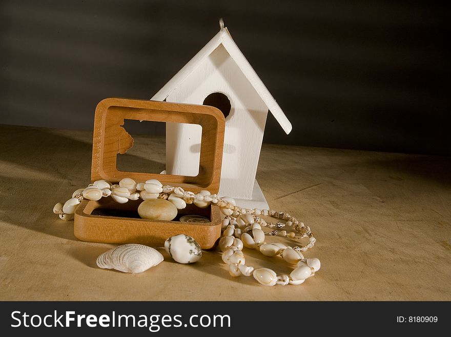 White bird house with wooden storage device for personal items such as shell necklace, sea shells, and rock and on a wooden surface, against a gray background with a window blind shadow. White bird house with wooden storage device for personal items such as shell necklace, sea shells, and rock and on a wooden surface, against a gray background with a window blind shadow.