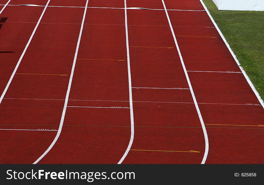 Straight red running track with white dividing lines part of grass visible. Straight red running track with white dividing lines part of grass visible