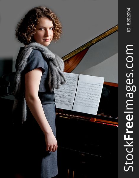 Young girl with serious look in gray dress and fur scarf near black piano with music notes standing turned right looking forward on grey backgorund. Young girl with serious look in gray dress and fur scarf near black piano with music notes standing turned right looking forward on grey backgorund