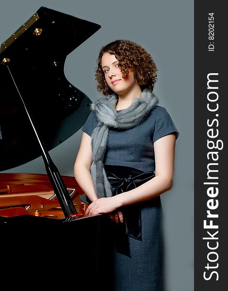 Young girl with curious look in gray dress and fur scarf near black piano  standing turned left with hands crossed appear looking forward on grey backgorund