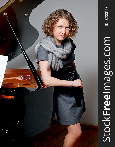 Young girl with curious look in gray dress and fur scarf near black piano standing tilted right supported looking forward smiling slaightly on grey backgorund