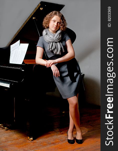 Young girl with curious look in gray dress and fur scarf near black piano standing turned left supported looking forward smiling slaightly on grey backgorund