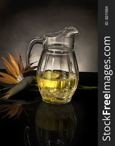 Glass pitcher filled with lemonade, next to a bird of paradise flower on a black reflective surface, against a gray background with a spot light. Glass pitcher filled with lemonade, next to a bird of paradise flower on a black reflective surface, against a gray background with a spot light.