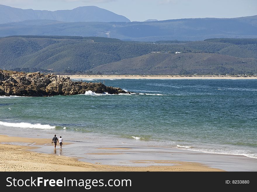 A middle-aged couple strolls along the beach with mountains in the distance and bright blue ocean. A middle-aged couple strolls along the beach with mountains in the distance and bright blue ocean.