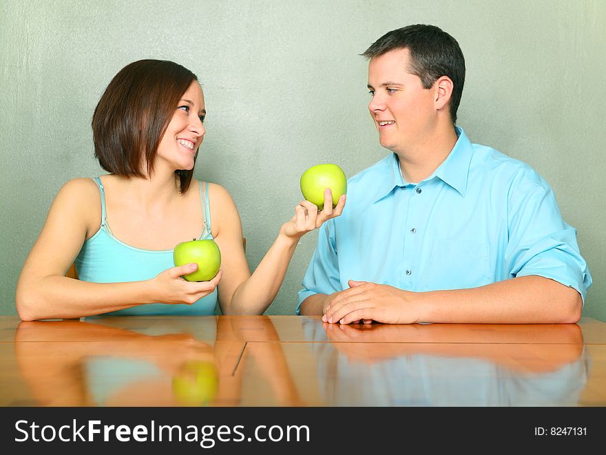 Beautiful young female caucasian offering green apple to her boyfriend or husband on kitchen table. Beautiful young female caucasian offering green apple to her boyfriend or husband on kitchen table