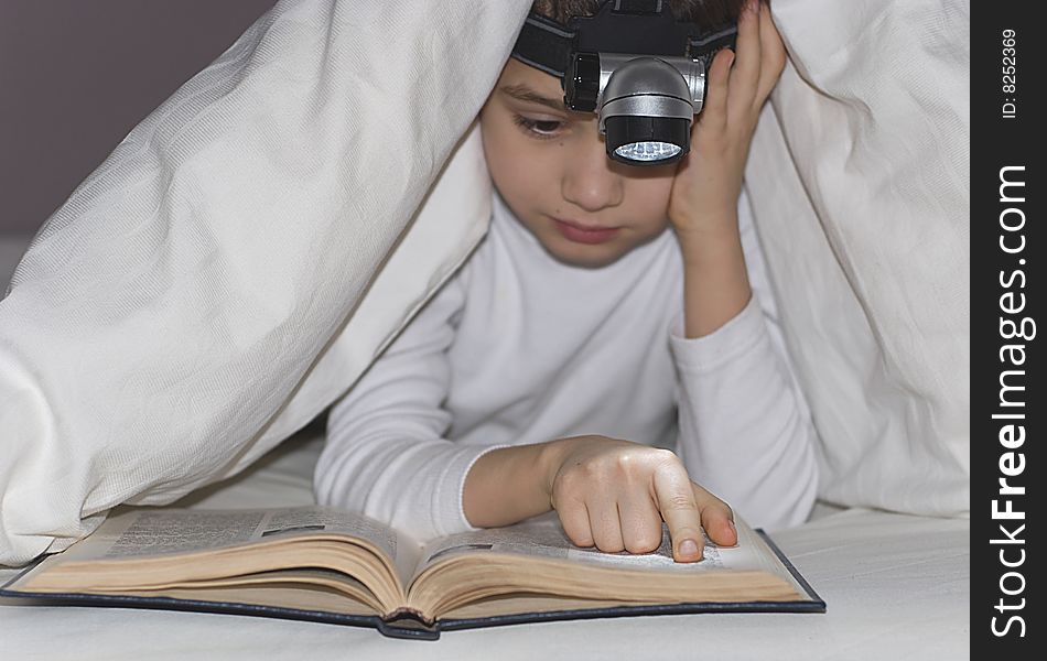 The boy has hidden under a blanket with a small lamp and reads the book. The boy has hidden under a blanket with a small lamp and reads the book