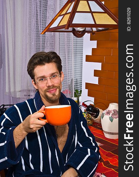 Young man wearing blue striped bathrobe sitting in his kitchen and drinking his breakfast tea. Young man wearing blue striped bathrobe sitting in his kitchen and drinking his breakfast tea