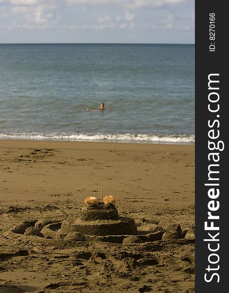 A sand castle in the forground and a soft focus person floating in the ocean in the background