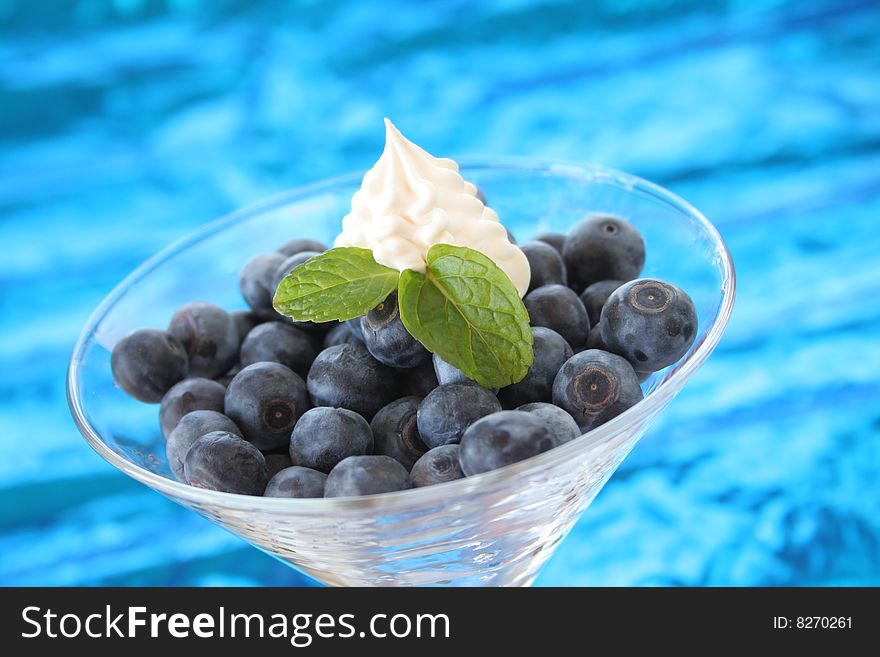 Blueberries with mint leaves and whip topping in a martini glass and shot on a metalic blue background. Blueberries with mint leaves and whip topping in a martini glass and shot on a metalic blue background.