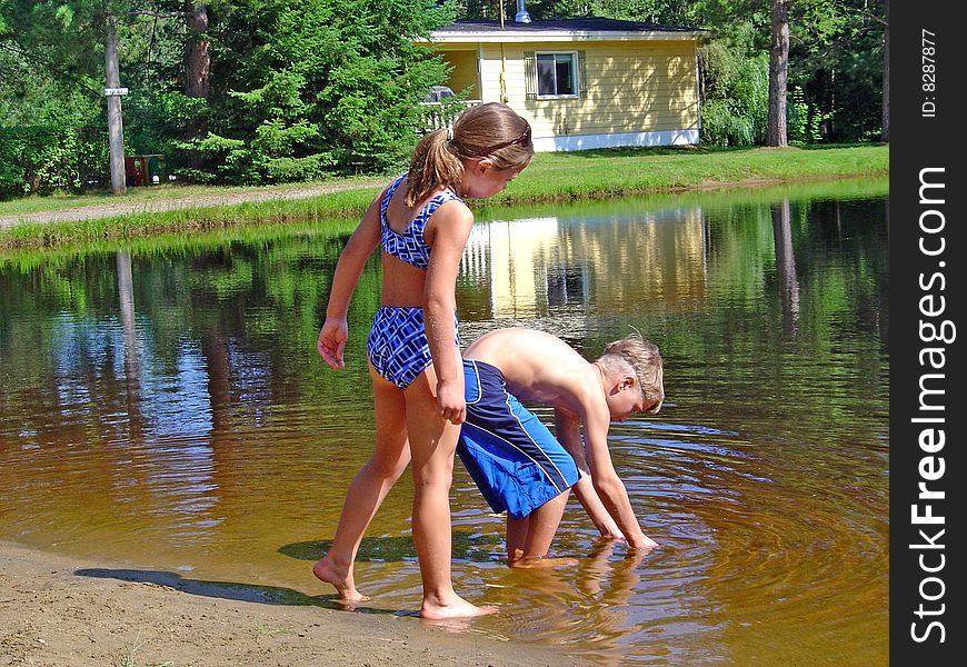 A boy is trying to catch some small fish in the lake while a girl looks on. A boy is trying to catch some small fish in the lake while a girl looks on