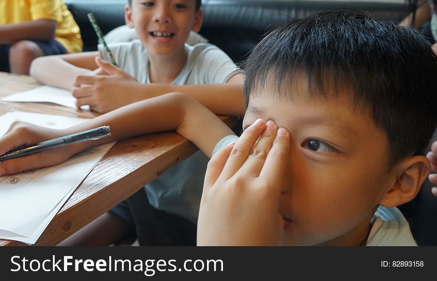 Young boy gesturing to forehead while doing schoolwork at table inside. Young boy gesturing to forehead while doing schoolwork at table inside.