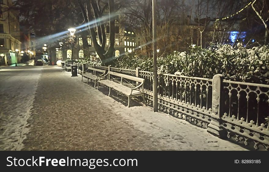 Snow covered fencing and bench on sidewalk and street illuminated in urban setting at night. Snow covered fencing and bench on sidewalk and street illuminated in urban setting at night.