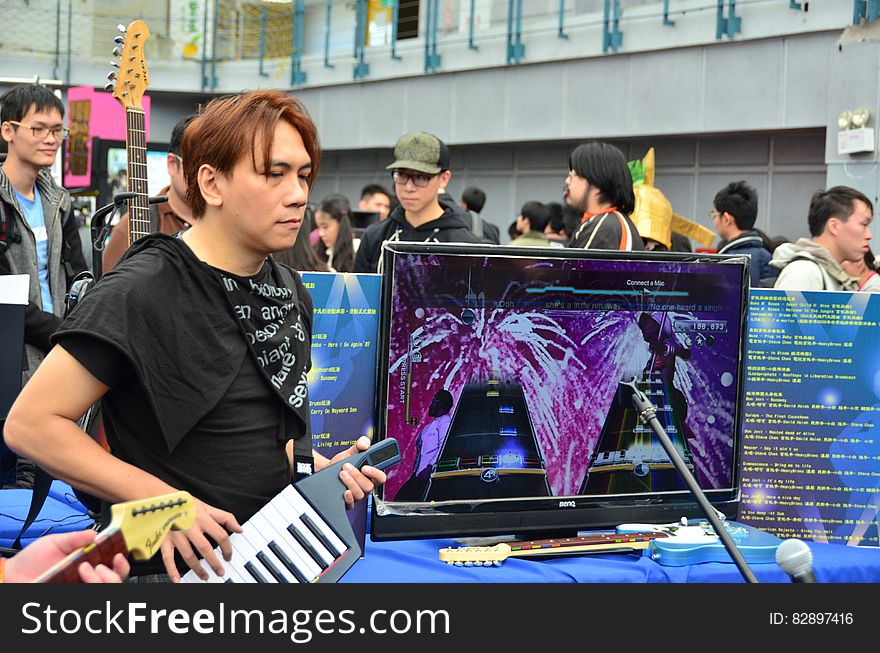 Young person playing a keyboard at a music fair next to a computer display screen with crowd of people in the background. Young person playing a keyboard at a music fair next to a computer display screen with crowd of people in the background.