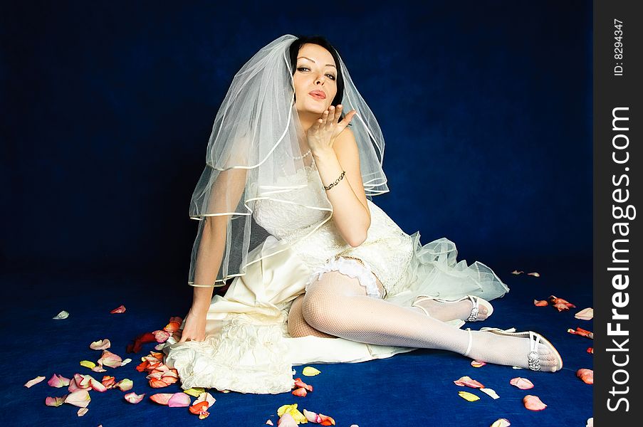 Studio portrait of a beautiful bride sitting on the floor covered with rose leaves and sending us an air kiss. Studio portrait of a beautiful bride sitting on the floor covered with rose leaves and sending us an air kiss