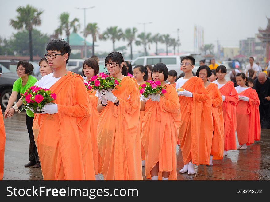 Children in orange robes holding flowers in outdoor religious procession.