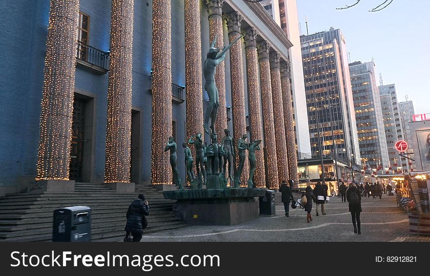 Pillars and statue outside modern urban architecture with crowds on sidewalk at twilight. Pillars and statue outside modern urban architecture with crowds on sidewalk at twilight.