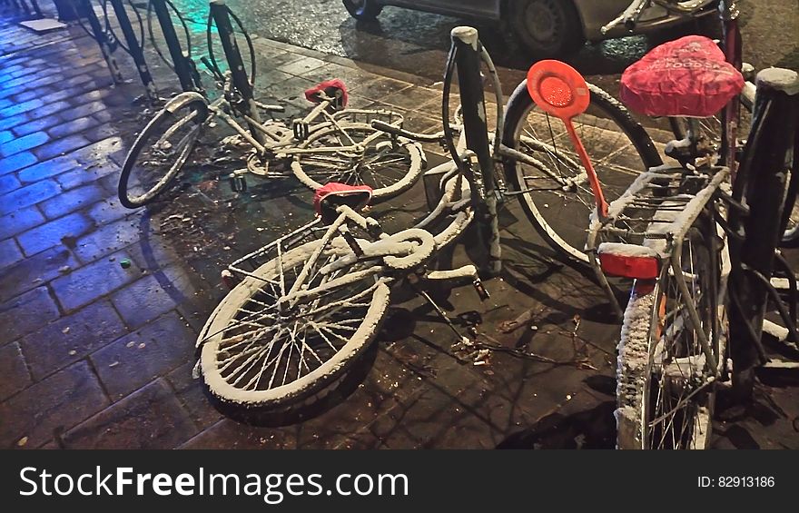 Bicycles untidily parked on the pavement at the roadside but chained up to stout fixed black posts. One bike has a red cover, rear carrier and light. Bicycles untidily parked on the pavement at the roadside but chained up to stout fixed black posts. One bike has a red cover, rear carrier and light.