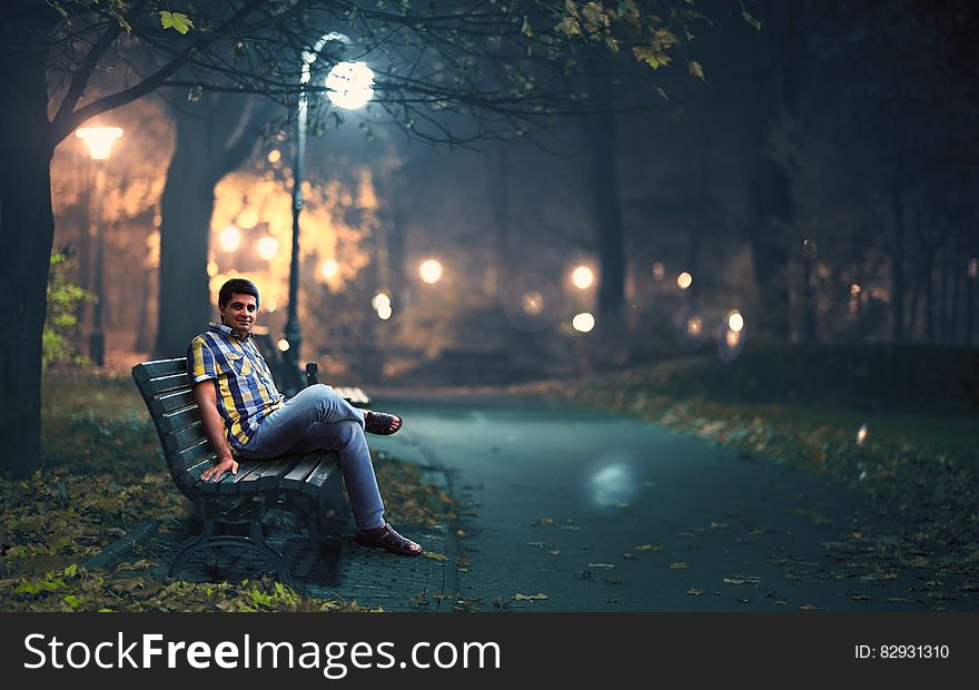 Man in Blue Denim Jeans Sitting Down by Wooden Bench Near Post Lamp Lighted
